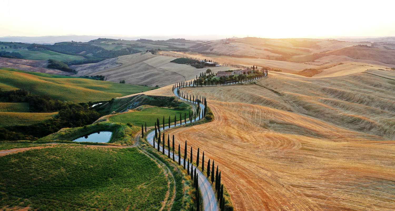 ASCIANO / ITALY - JULY 3 2019: Aerial view of Tuscany landscape and Asciano villa, curved road with cypress trees, agricultural field around at summer sunset. Siena, Italy.